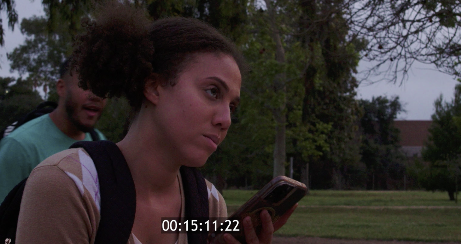 Performance still: Ashlyn outdoors with a phone and backpack, a scene partner just behind her.